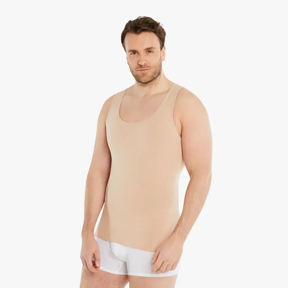 Ein selbstbewusster Mann trägt ein hautfarbenes Ärmelloses Kompressions-Unterhemd und schaut lächelnd in die Kamera. Das Unterhemd bietet einen starken Bauch-weg Effekt und besteht aus hochwertiger Baumwolle für Tragekomfort. Seine Arme sind entspannt an der Seite. #farbe_nude