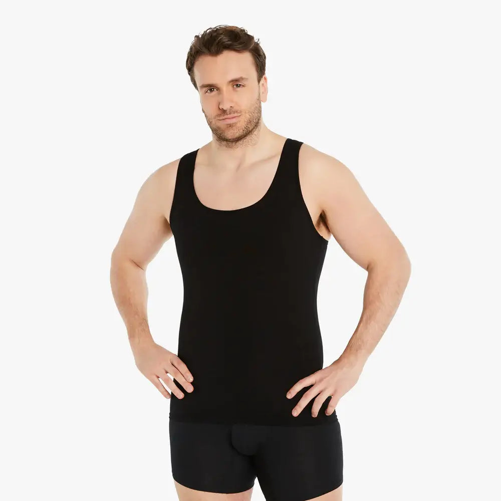 Ein selbstbewusster Mann mit einem schwarzen Ärmellosen Kompressions-Unterhemd schaut lächelnd in die Kamera, während er die Arme in die Hüften stemmt. Das Unterhemd bietet einen starken Bauch-weg Effekt und besteht aus hochwertiger Baumwolle für Tragekomfort. #farbe_schwarz