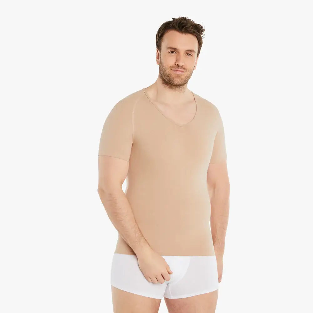 Ein selbstbewusster Mann lächelt in die Kamera, trägt ein hautfarbenes Kurzarm Shapewear Kompressions-Unterhemd. Seine Hand ruht leicht vor dem Körper. Das Unterhemd bietet einen starken Bauch-weg Effekt, besteht aus hochwertiger Baumwolle und garantiert Tragekomfort. #farbe_nude