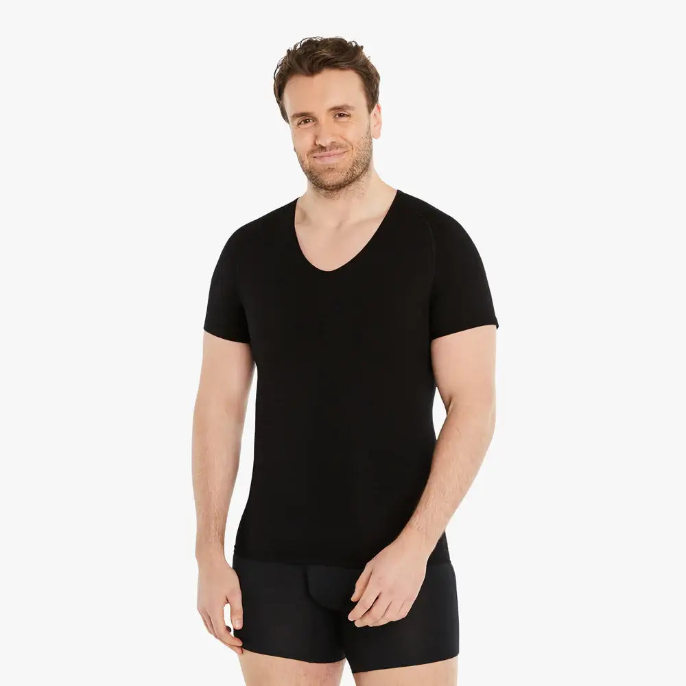 Ein selbstbewusster Mann lächelt in die Kamera, trägt ein schwarzes Kurzarm Shapewear Kompressions-Unterhemd. Seine Hand ruht leicht vor dem Körper. Das Unterhemd bietet einen starken Bauch-weg Effekt, besteht aus hochwertiger Baumwolle und garantiert Tragekomfort. #farbe_schwarz