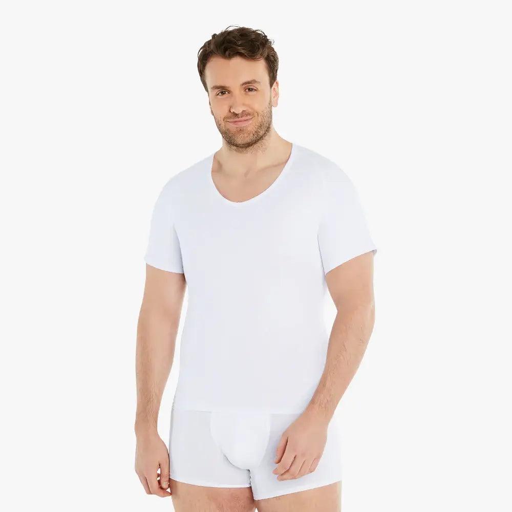 Selbstbewusster Mann lächelt in die Kamera, während er ein weißes Kurzarm Shapewear Kompressions-Unterhemd trägt und entspannt die Arme an der Seite hält. Das Unterhemd bietet einen starken Bauch-weg-Effekt, besteht aus hochwertiger Baumwolle und garantiert hohen Tragekomfort. #farbe_weiß
