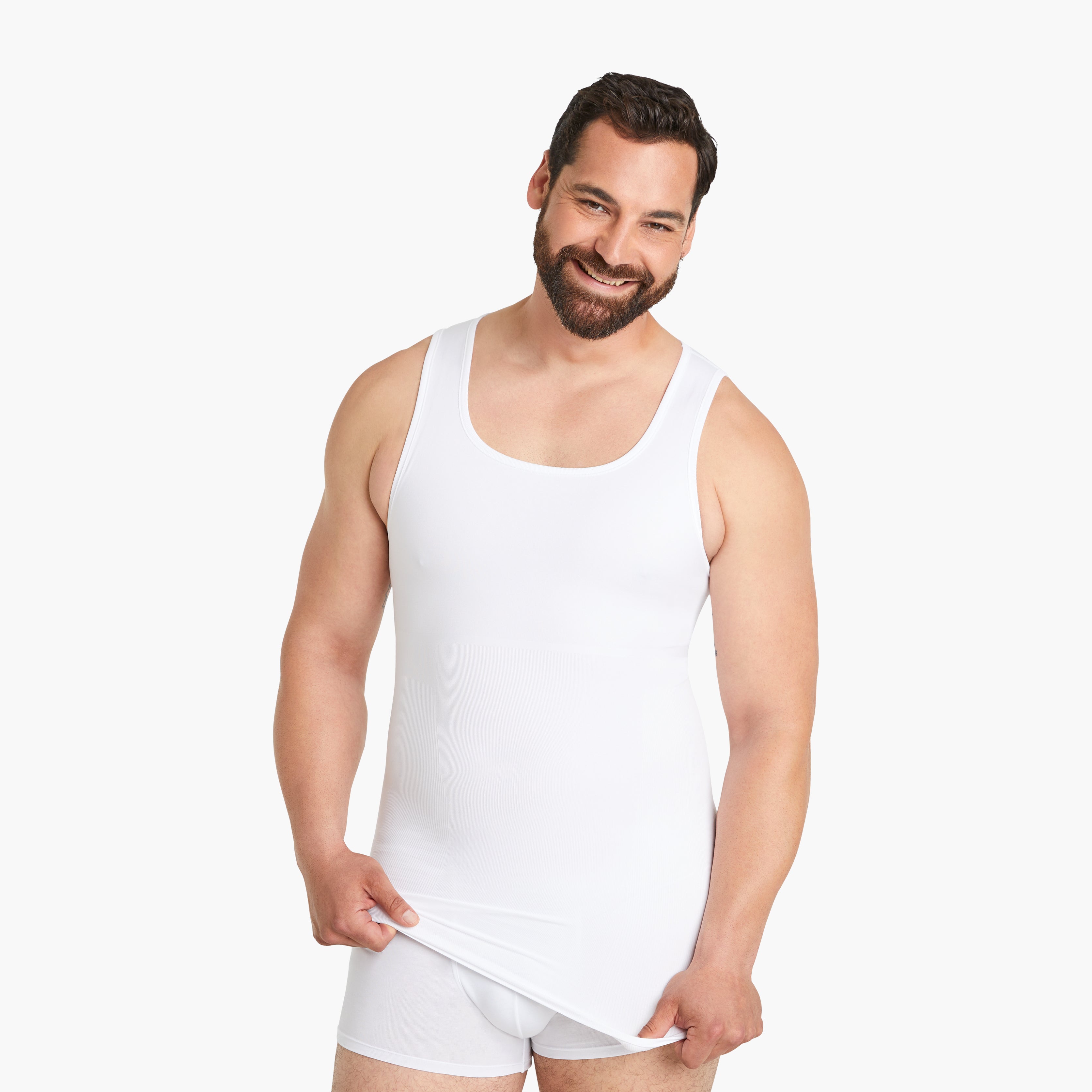 Auf dem Bild trägt ein Mann ein weißes Seamless Shapewear Tank-Top für Herren. Mit einem lächelnden Gesicht zieht er das Shirt nach unten, während er in die Kamera schaut. Dieses Tank-Top bietet einen Bauch-weg Effekt dank innovativer Seamless Technologie und verspricht hohen Tragekomfort. #farbe_weiß