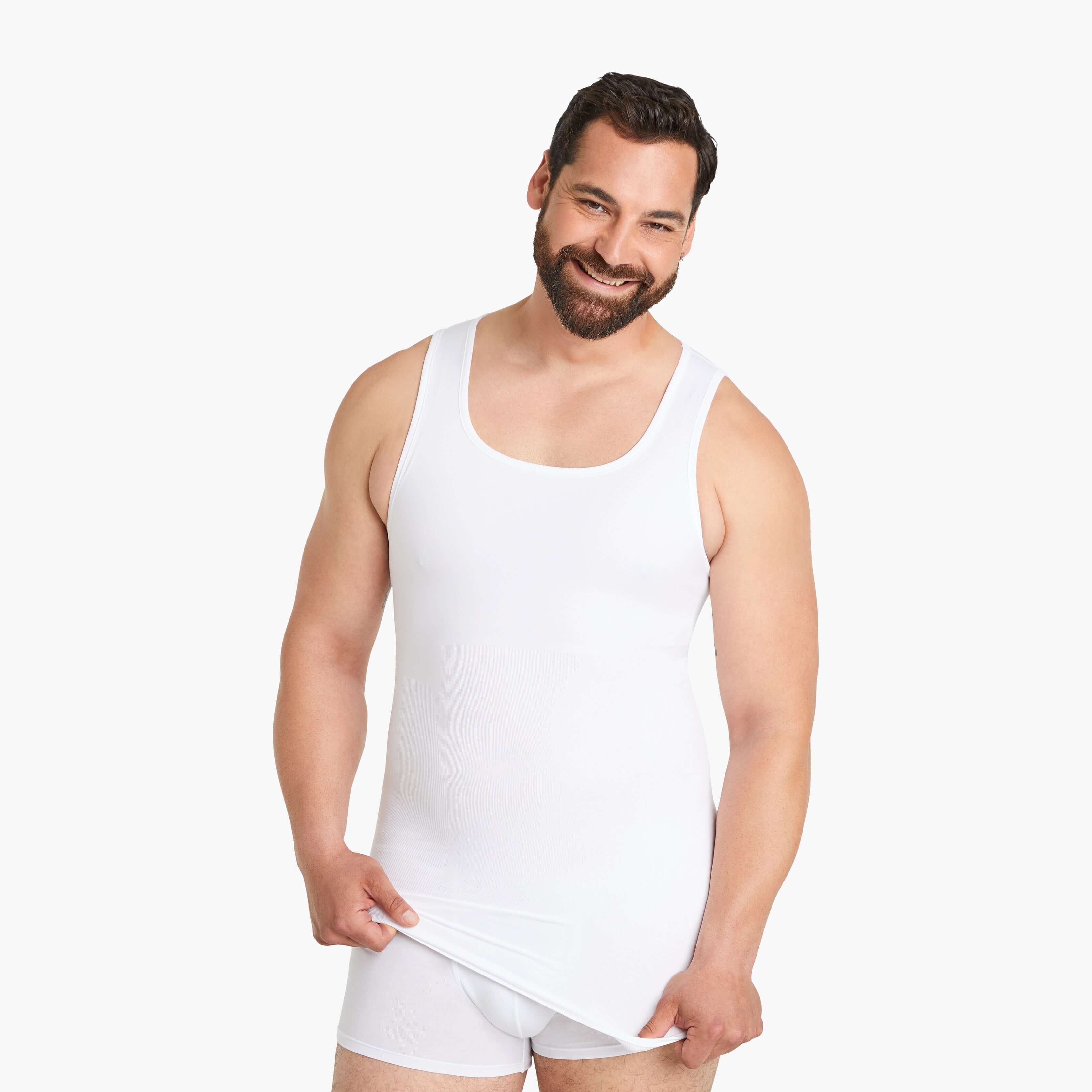 Mann trägt lächelnd ein weißes Seamless Shapewear Tank-Top und zieht es nach unten, während er in die Kamera schaut. Das Tank-Top bietet einen Bauch-weg-Effekt durch innovative Seamless-Technologie und garantiert hohen Tragekomfort. #farbe_weiß